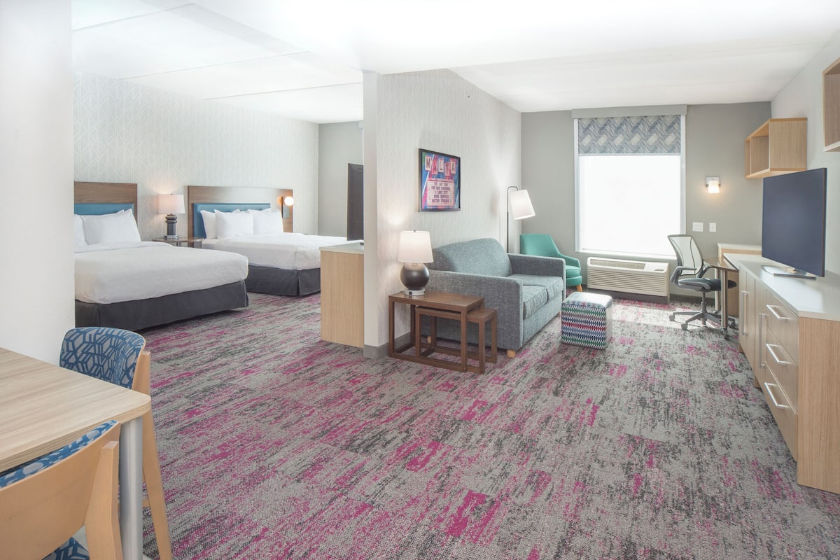 A hotel room with two beds, a desk, a chair, a TV, and a window with blinds.