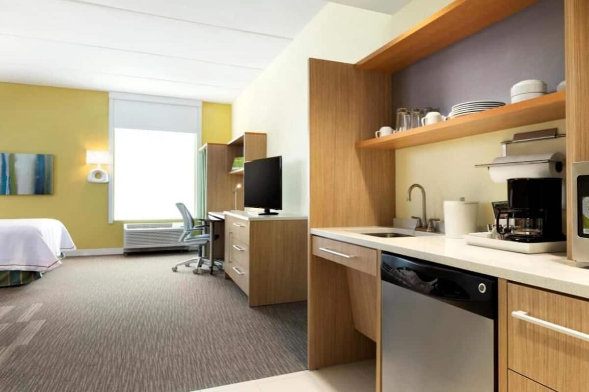 A modern hotel room with a kitchenette, a bed, a desk, and a television.