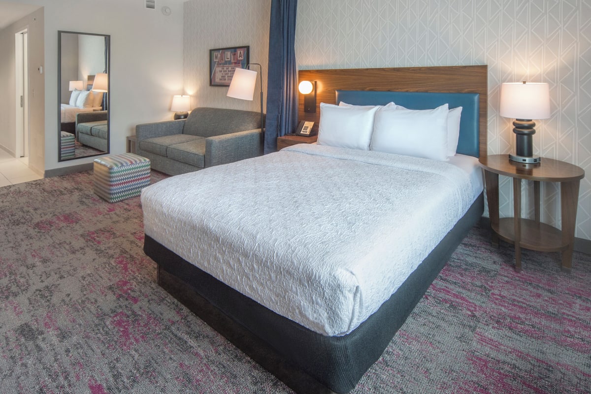 A hotel room with a large bed, a sofa, a small table, a mirror, and a nightstand with a lamp.