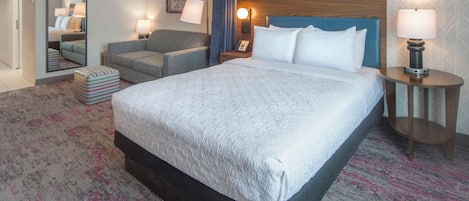 A hotel room with a large bed, a sofa, a small table, a mirror, and a nightstand with a lamp.