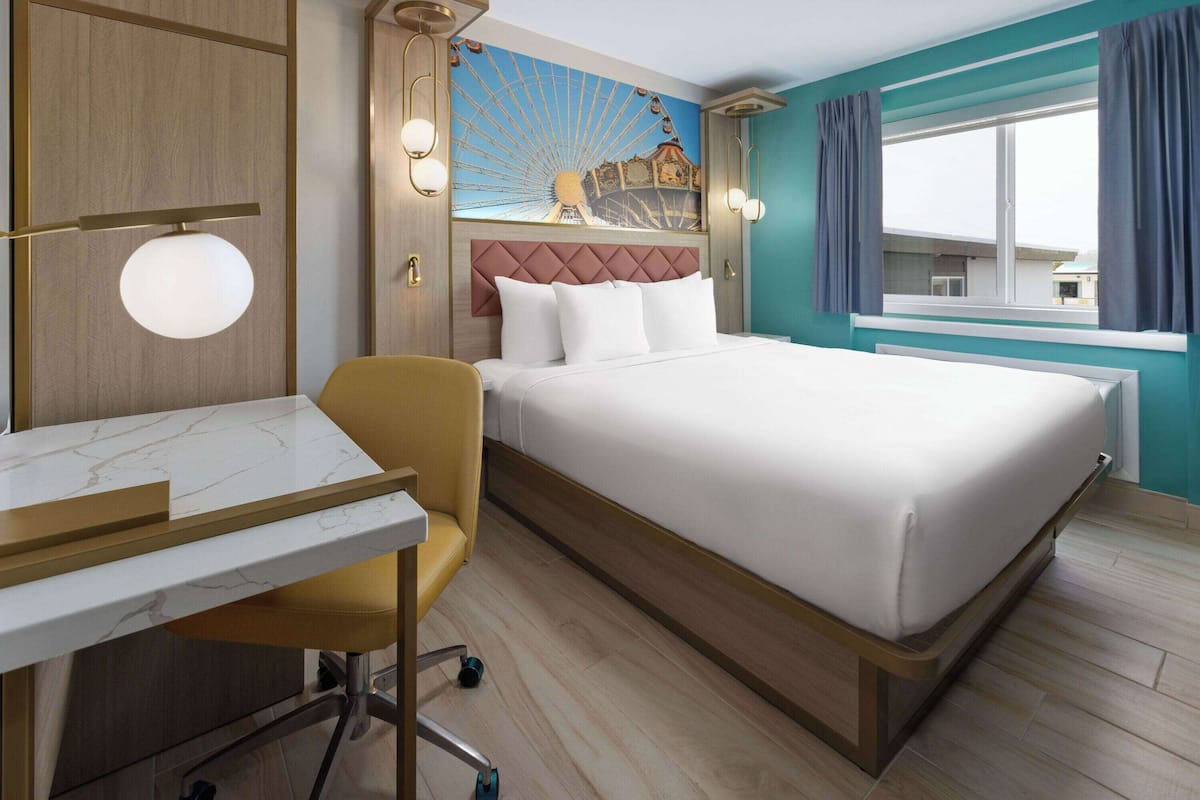 Habitación de hotel con cama, escritorio, silla y un ventanal grande, además de un mural de noria.
