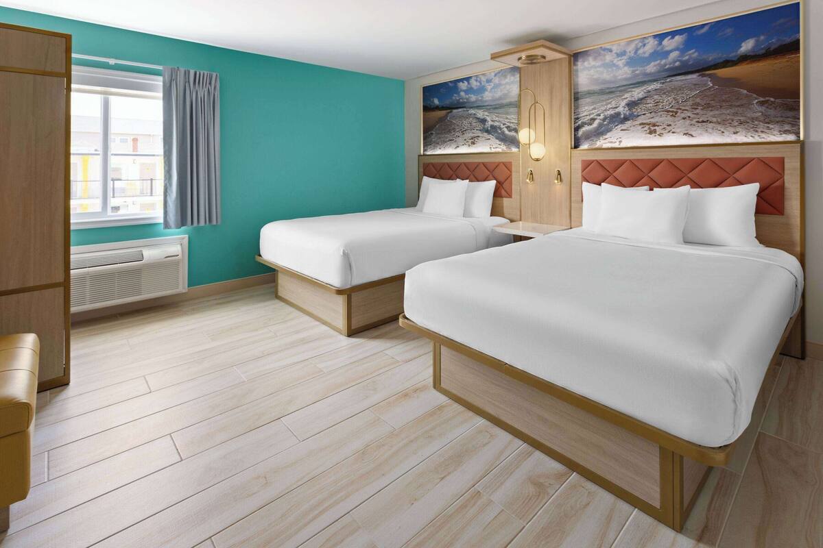 Habitación de hotel con dos camas, un ventanal con cortinas, un calentador en la pared y un cuadro con motivos playeros sobre las camas.