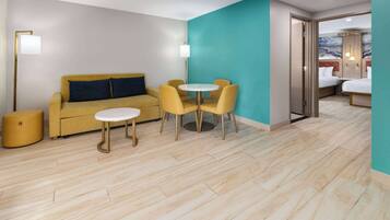 A modern hotel room with a sofa, a round table with chairs, a bed, and a view of the sea.