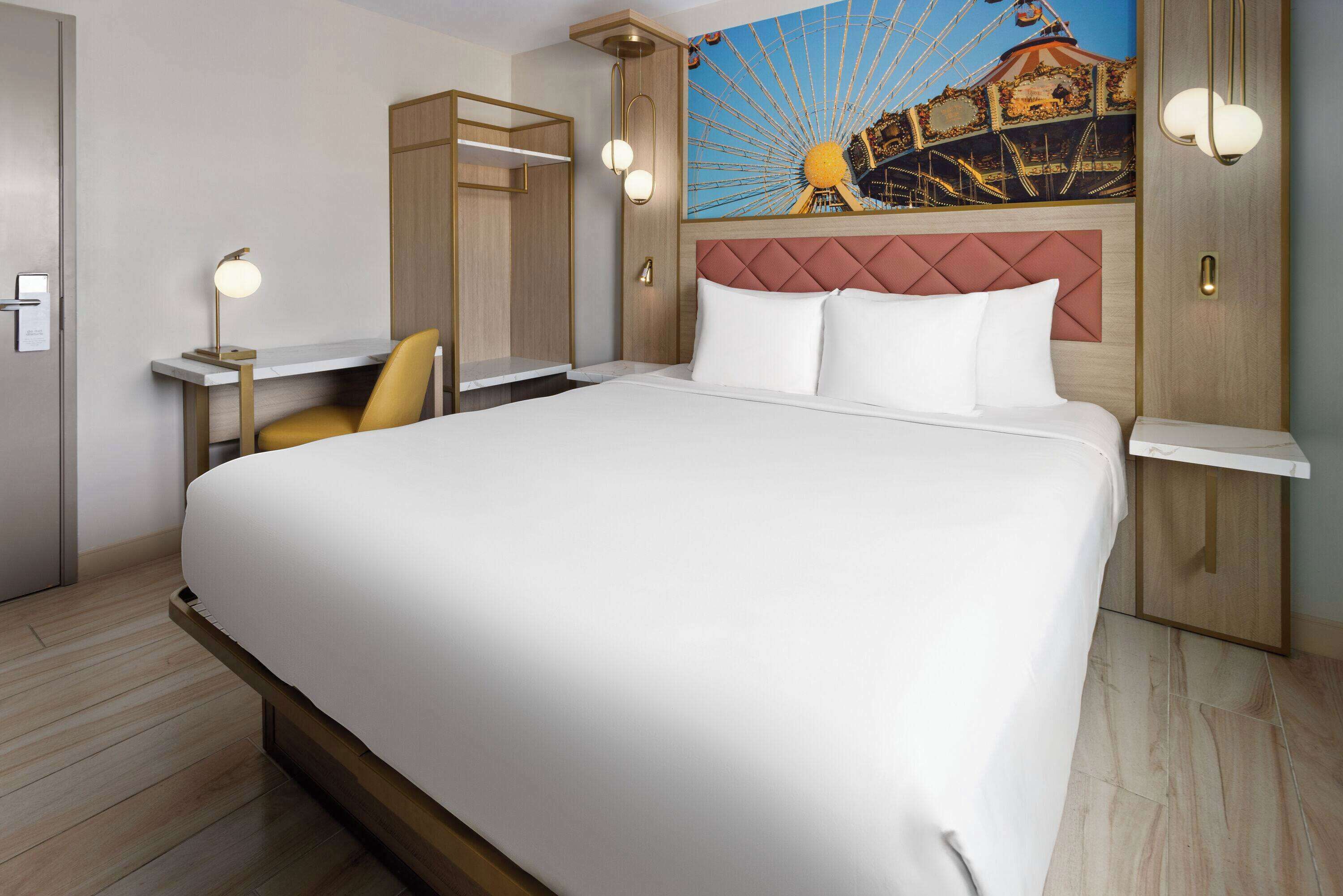 Quarto de hotel com uma cama grande, uma escrivaninha, uma cadeira e um quadro de uma roda gigante.