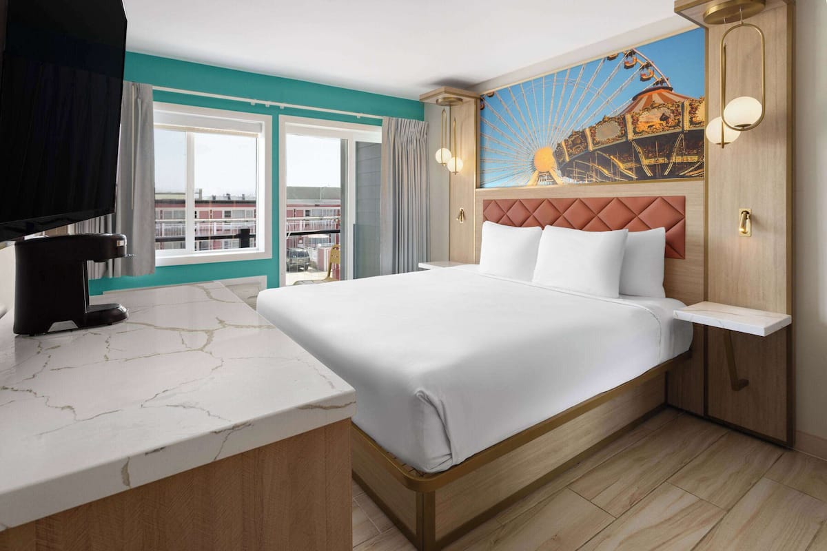 Habitación de hotel con una cama grande, una mesita de noche de mármol, un televisor montado, un cuadro de una noria y vistas a la ciudad a través de un ventanal amplio.