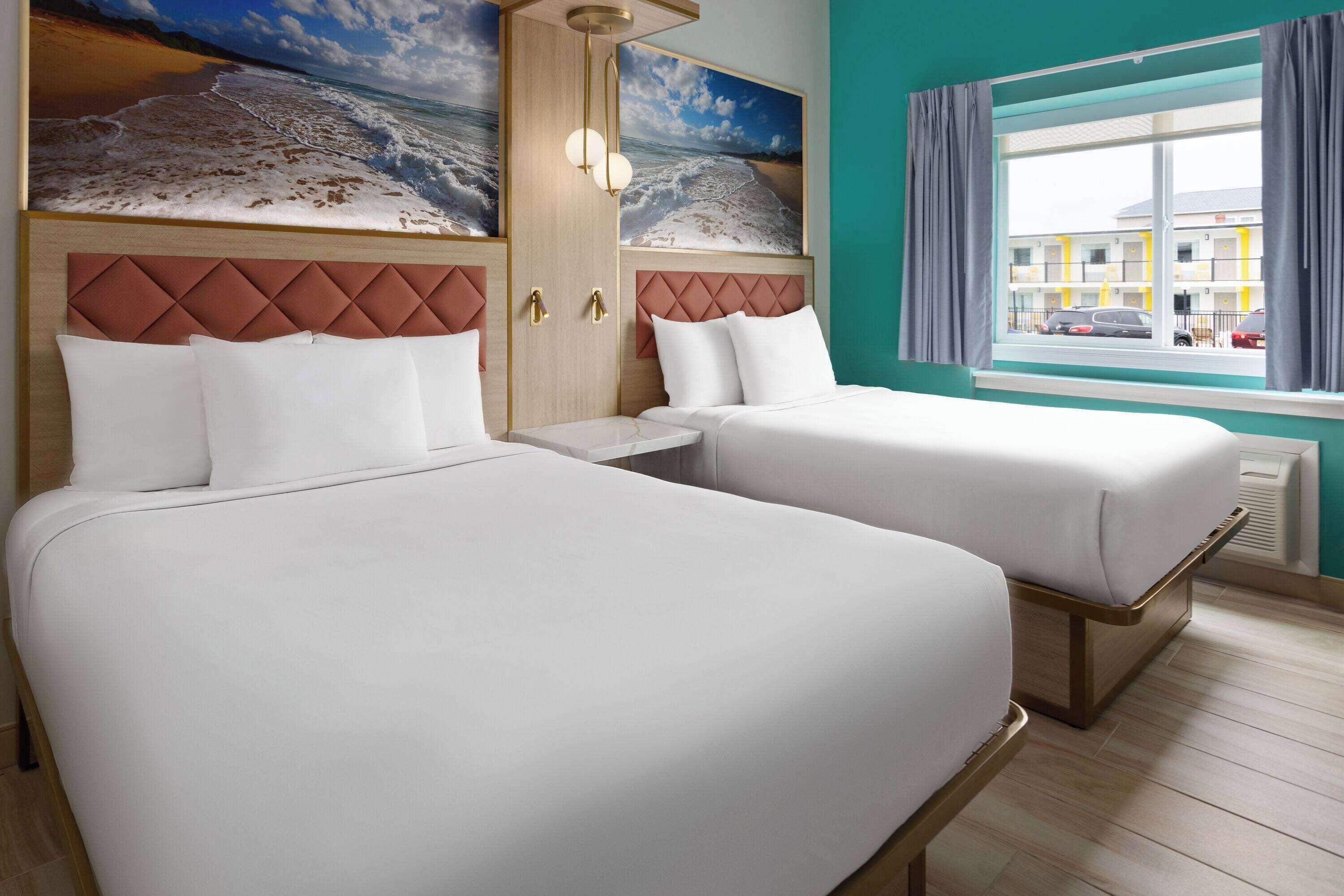 A hotel room with two beds, a large window with curtains, a view of a building outside, and a beach scene on the wall.