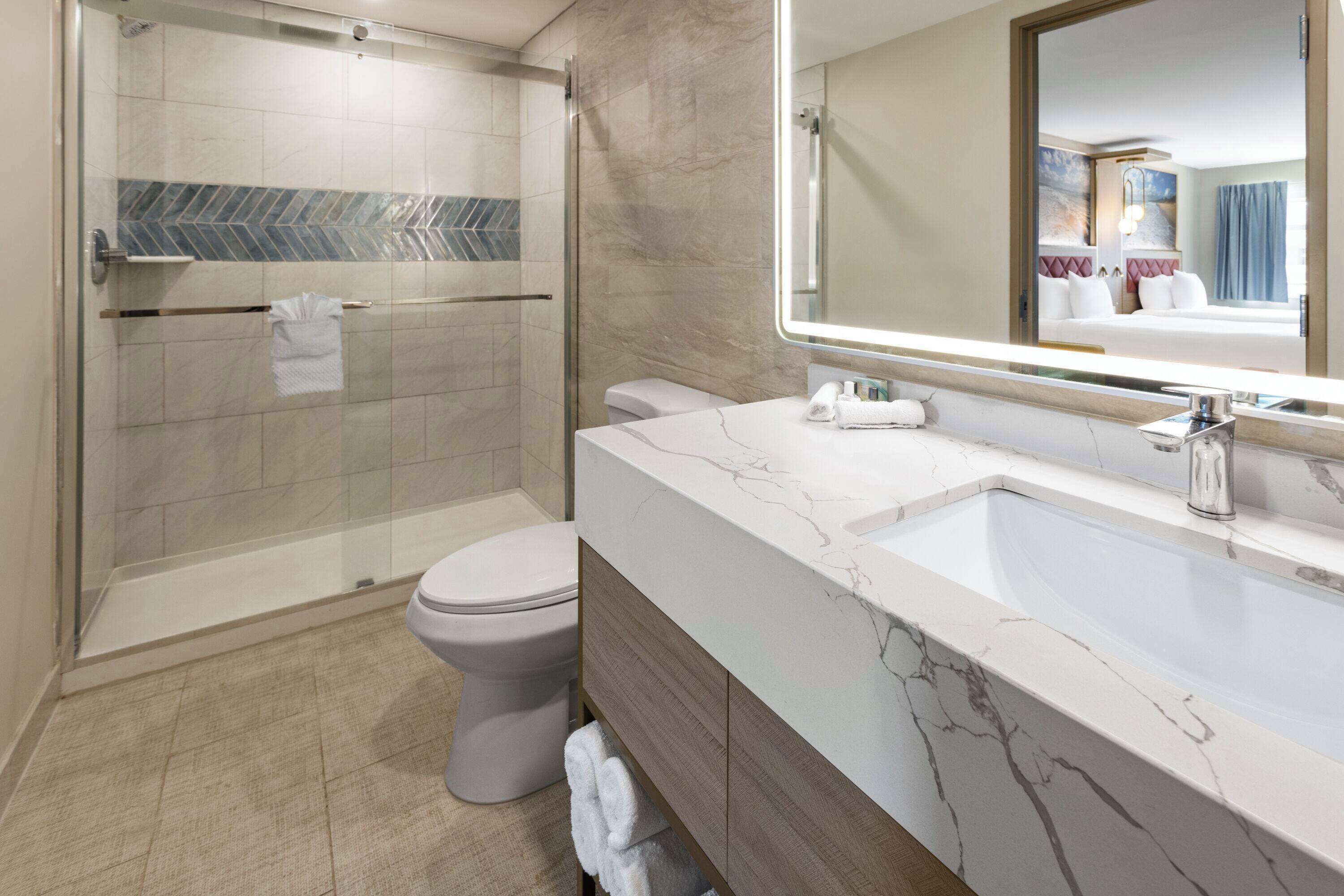 Une salle de bain moderne avec une douche à parois de verre, des toilettes blanches et un meuble-lavabo en marbre agrémenté d’un grand miroir.