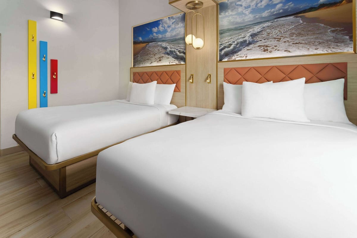 Una habitación de hotel con dos camas, un cabecero de madera, un cuadro de playa y una lámpara fijada a la pared.
