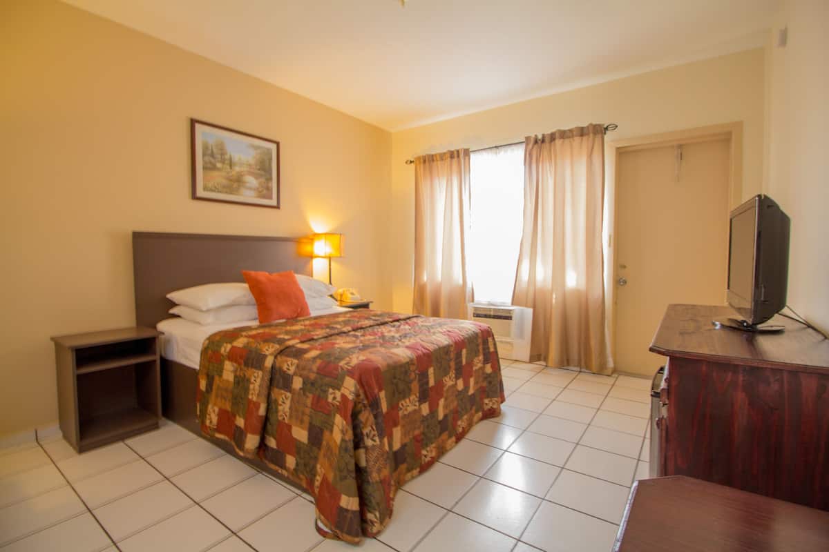 Une chambre d’hôtel avec un lit, une table de chevet, un bureau avec une télévision et une fenêtre avec des rideaux.