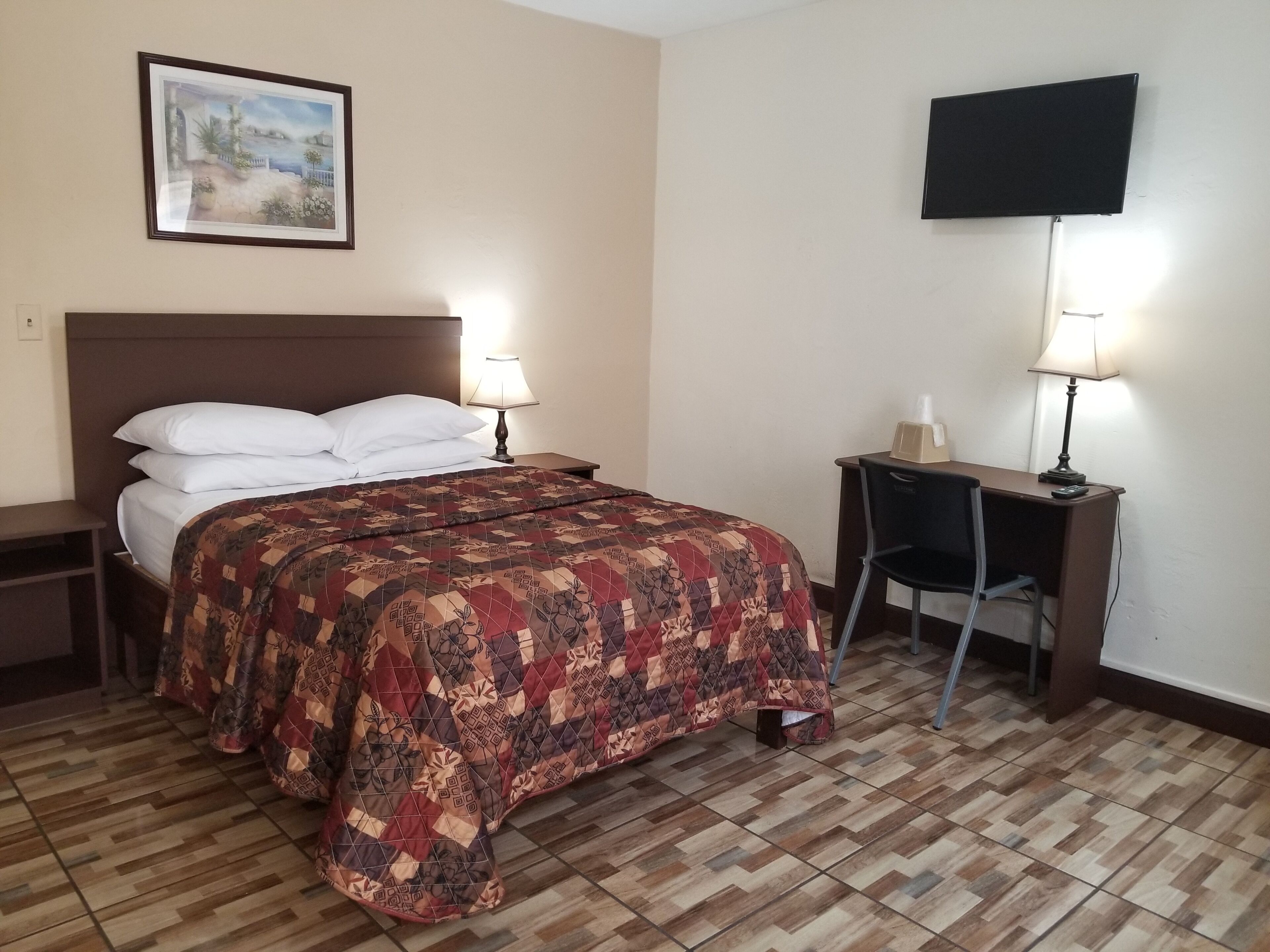 A hotel room with a bed, two bedside lamps, a desk with a chair, a television on the wall, and a framed picture above the bed.