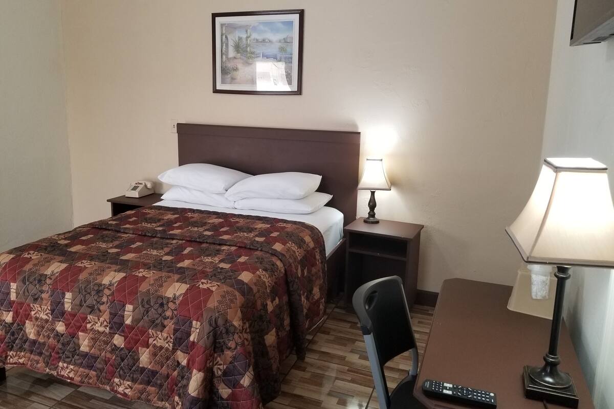 Une chambre d’hôtel avec un lit, une table de chevet avec une lampe, un bureau avec une télécommande et un tableau encadré au mur.