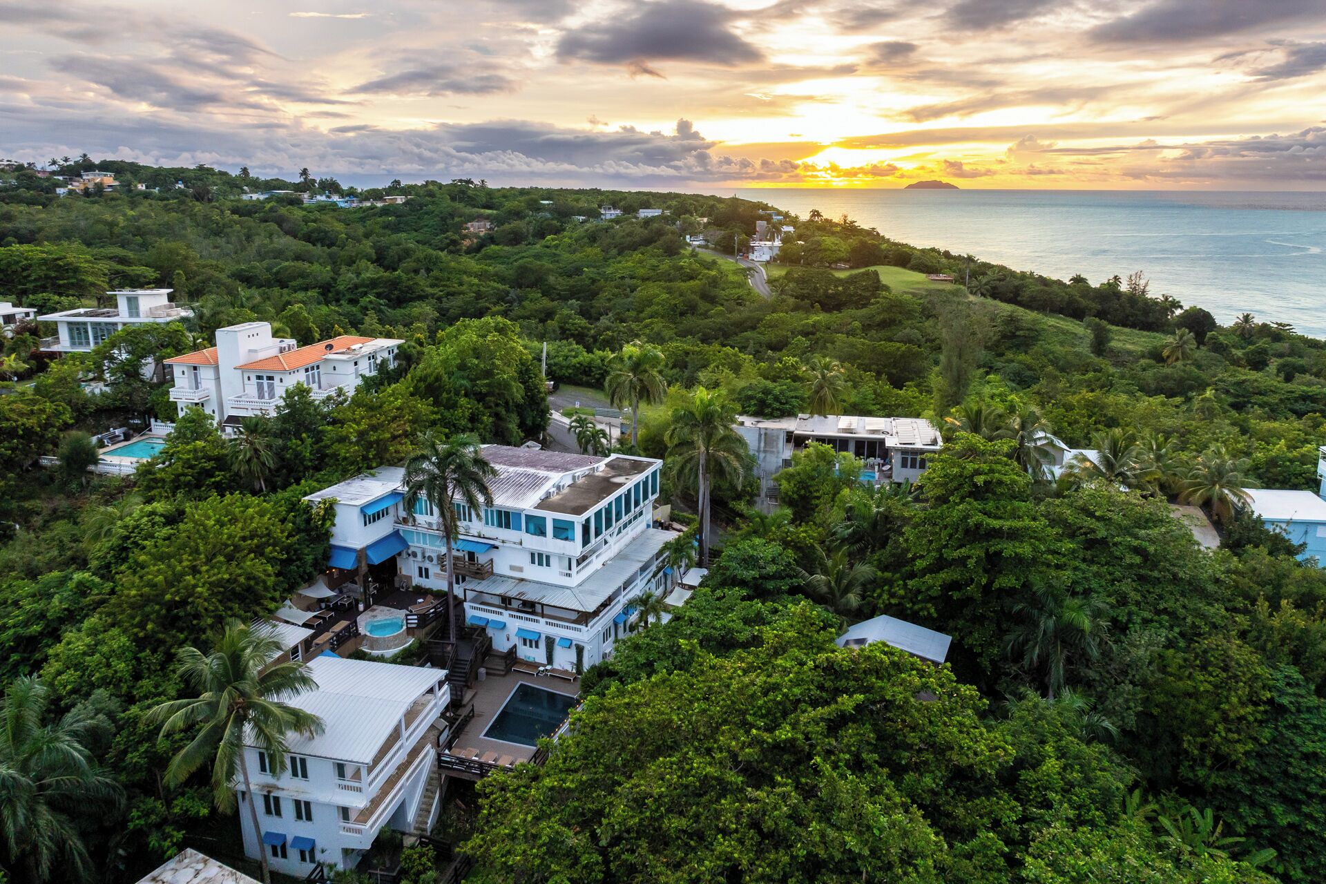 A coastal residential area with houses surrounded by lush greenery and a view of the sea at sunset.