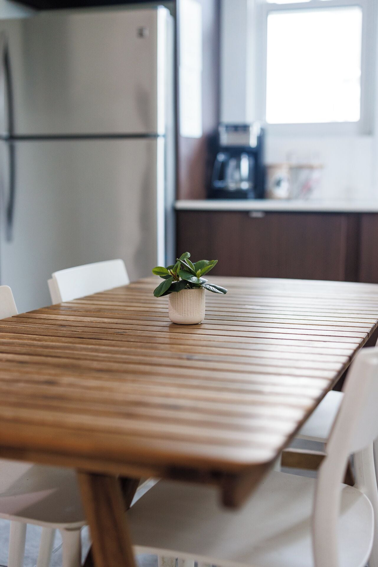 A wooden table with a potted plant in the center, surrounded by white chairs in a kitchen setting.