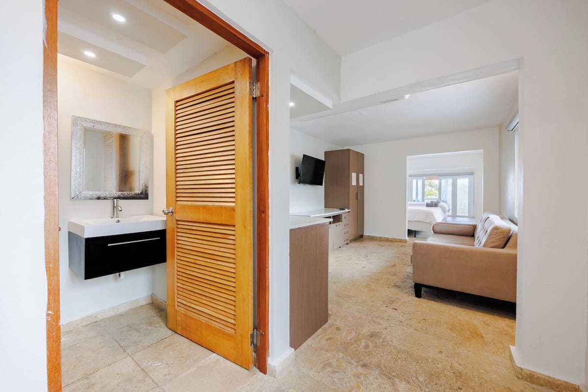 A modern interior with a wooden door, a bathroom with a mirror, a kitchenette, and a living area with a sofa.