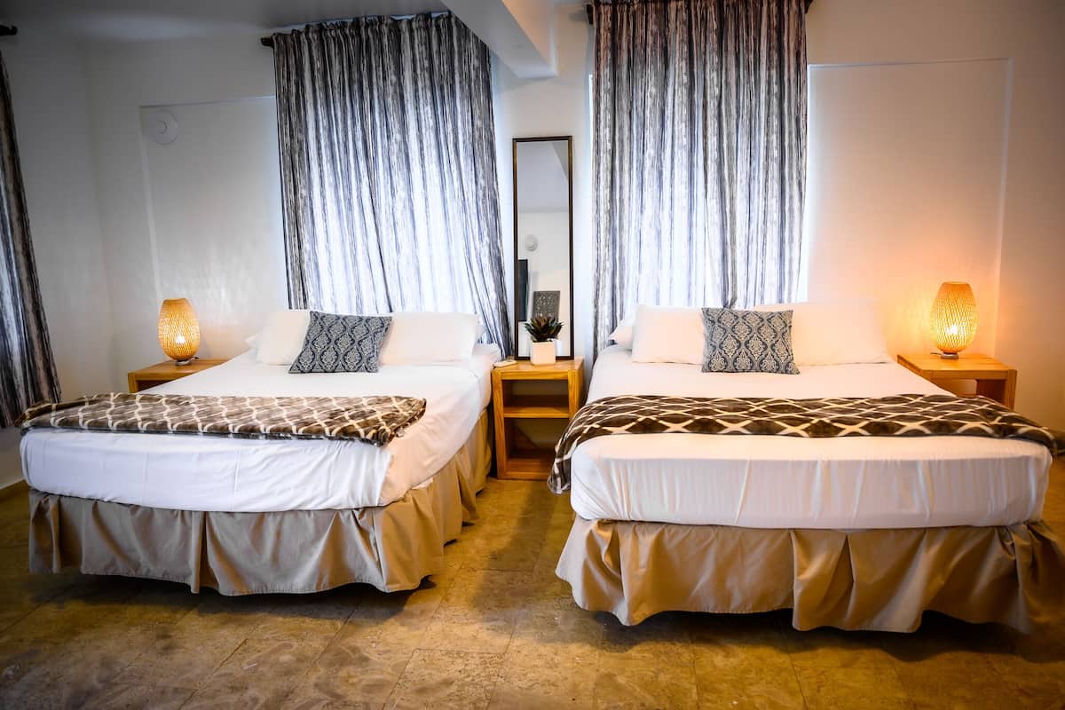 A hotel room with two single beds, each with a patterned bedspread and matching pillows. There are bedside tables with lamps, a small mirror, and a potted plant. The room has a neutral color scheme and a large mirror on the wall.