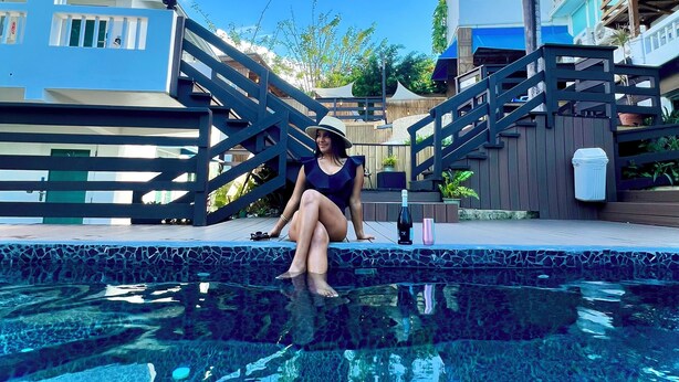 A person sitting by a swimming pool with a clear blue sky and palm trees in the background.