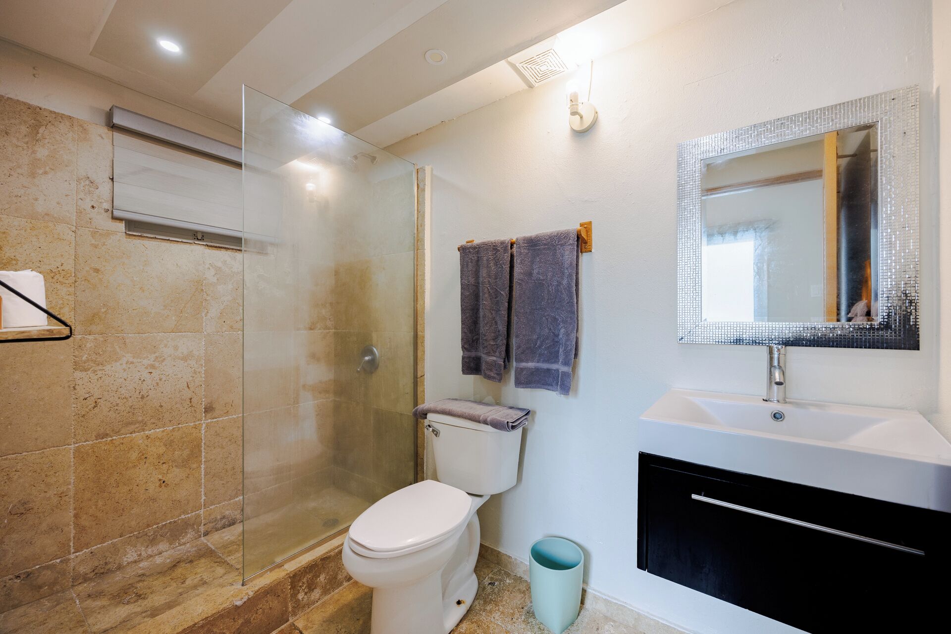 A bathroom with a glass-enclosed shower, a toilet, a sink with a mirror, and a trash bin.
