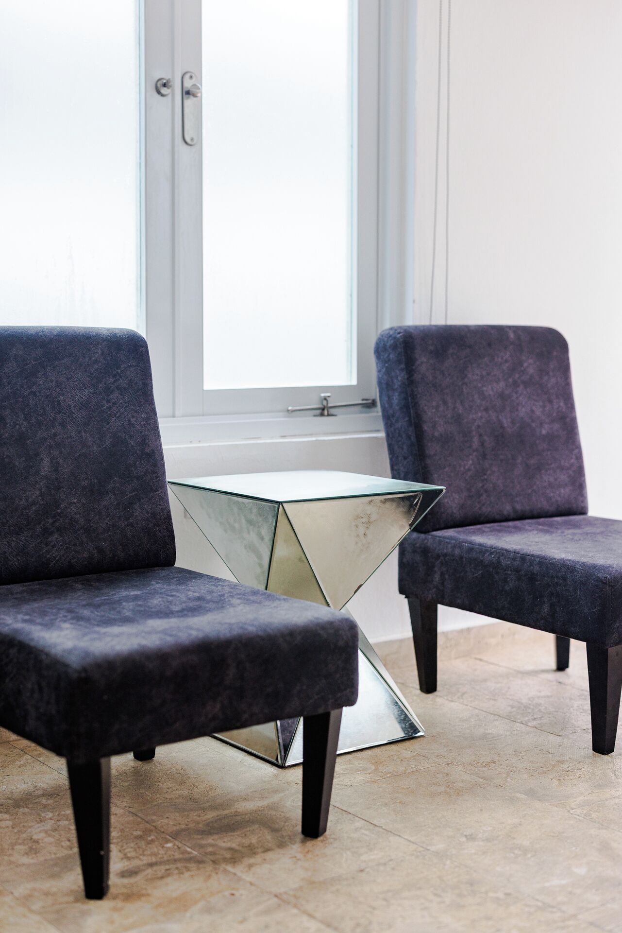 Two modern armchairs with a glass table in between, positioned in a room with a large window and a door.