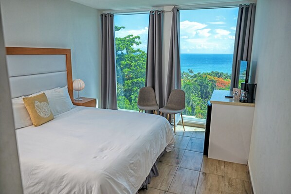 A hotel room with a bed, a desk, two chairs, and a view of the ocean. - Serenity Rincon Guesthouse (Rincon)
