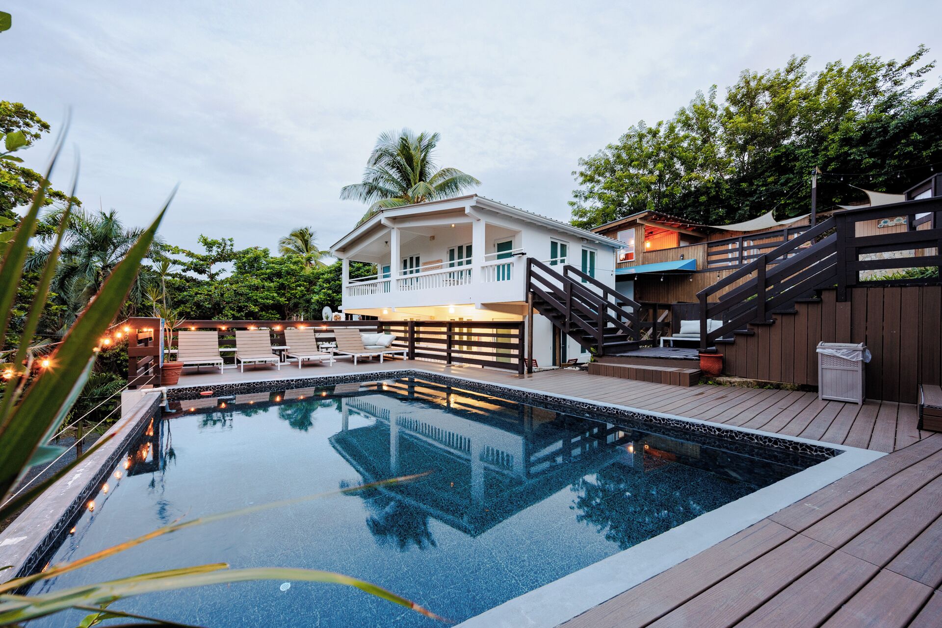 A pool area with a wooden deck, lounge chairs, and a white building with a balcony.