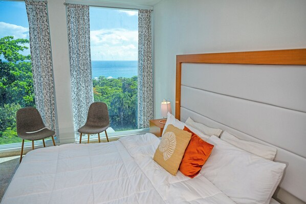 Habitación de hotel con una cama grande, dos sillas, una mesita de noche con lámpara y vistas al océano y la vegetación.