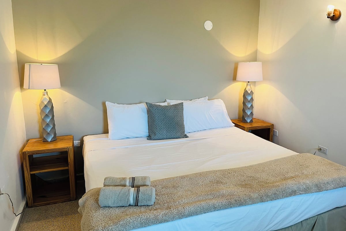 A neatly made bed with white and gray linens, two bedside lamps, a wooden nightstand, and a folded towel on the bed.