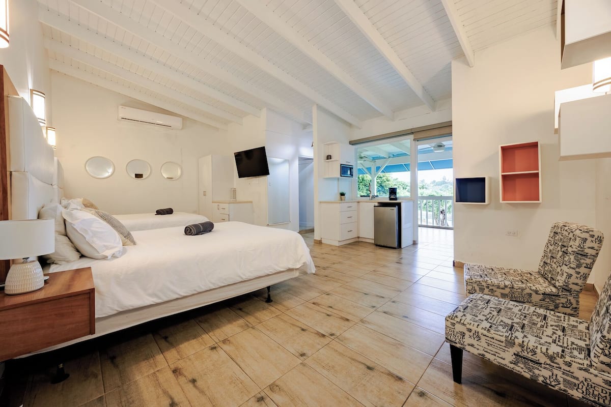 A hotel room with a large bed, a desk, a TV, a kitchenette, and a balcony with a view.