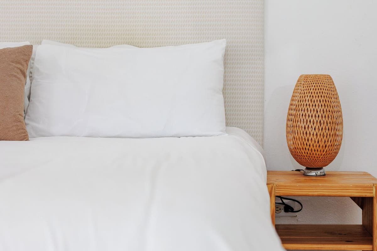 A neatly made bed with white bedding and a brown pillow, a woven lamp on a wooden nightstand, and a textured wall art piece above the bed.