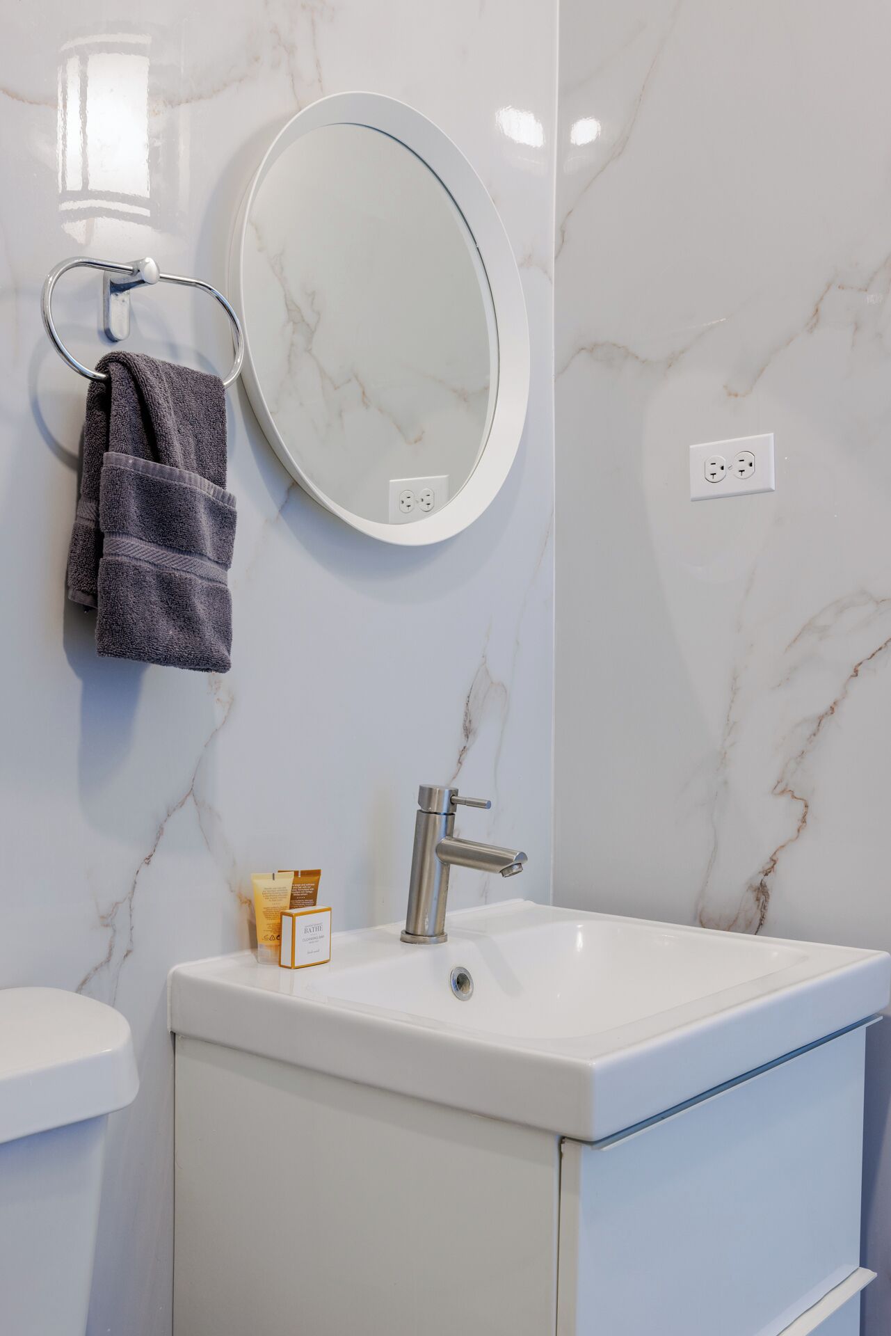 Un baño con lavamanos de mármol blanco, un espejo redondo, un perchero para toallas y una pequeña botella de productos de baño.