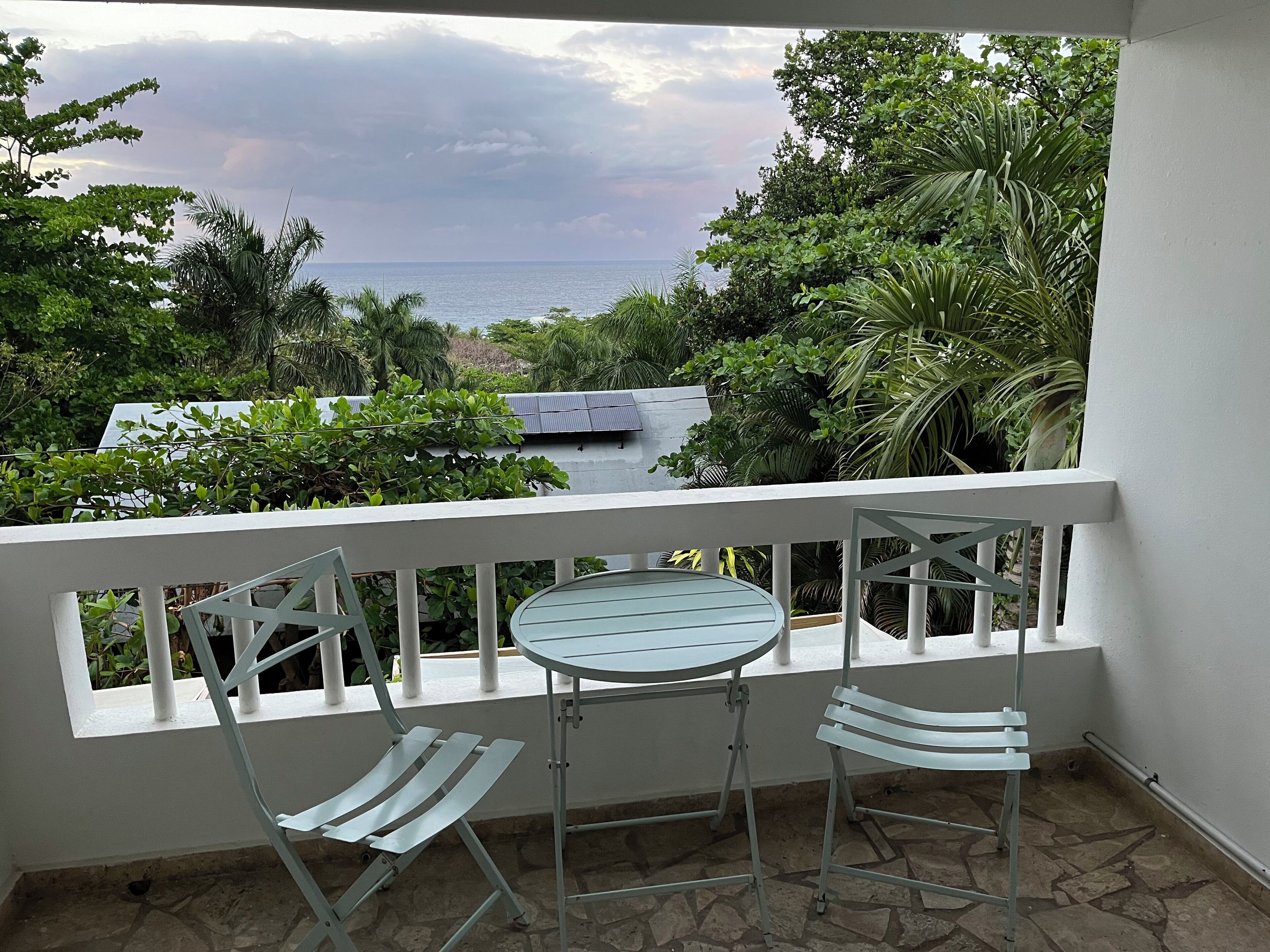 Un balcón con una mesa y dos sillas, con vista a un jardín exuberante y el mar.