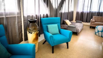 A living room with a blue armchair, a grey sofa, and a beige sofa. There is a large vase on a table and a window with curtains.