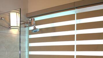 A shower with a glass door, a visible showerhead, and horizontal blinds partially covering the window.