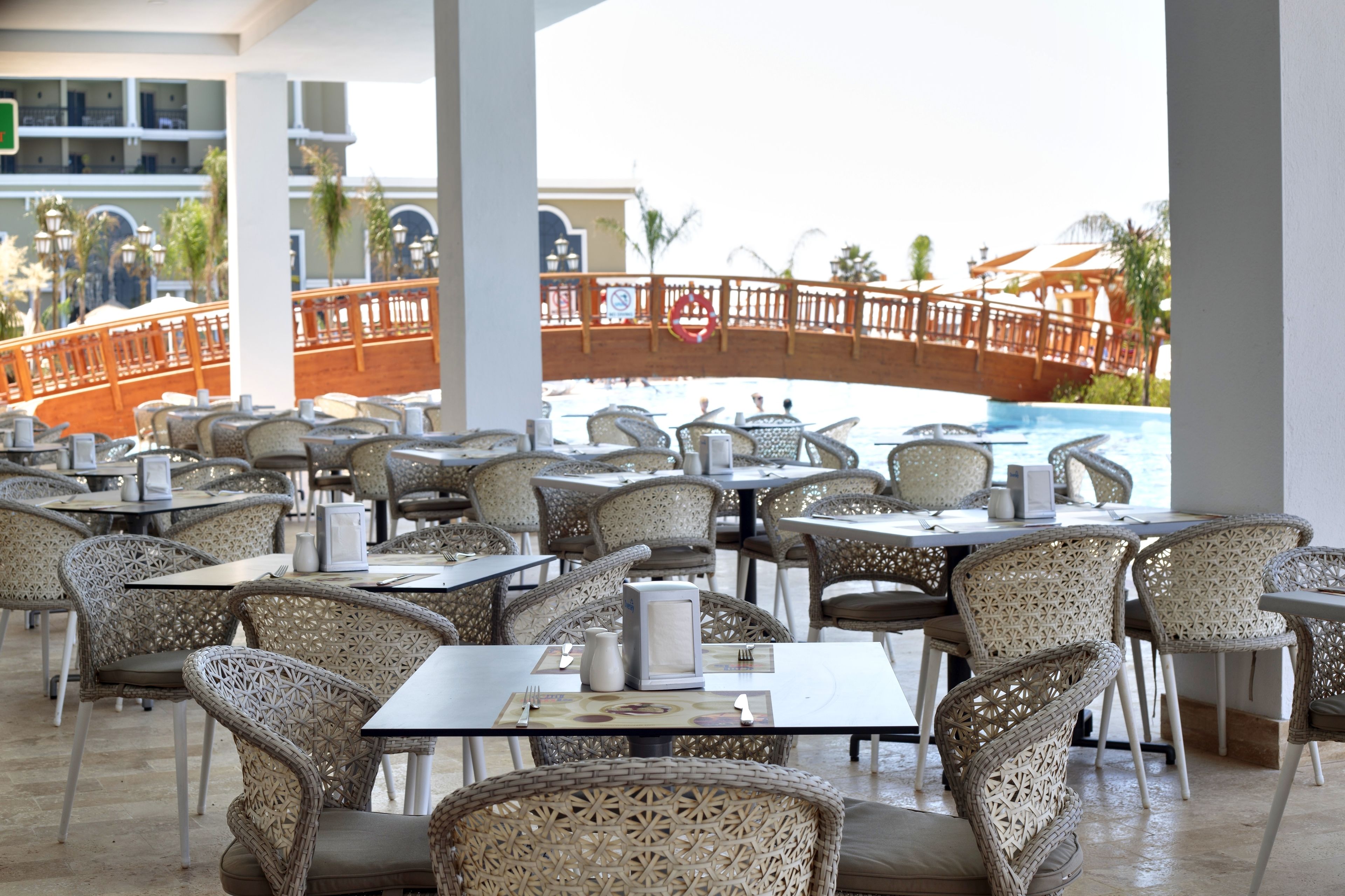 An outdoor dining area with wicker chairs and tables, a view of a bridge, and a pool in the background.