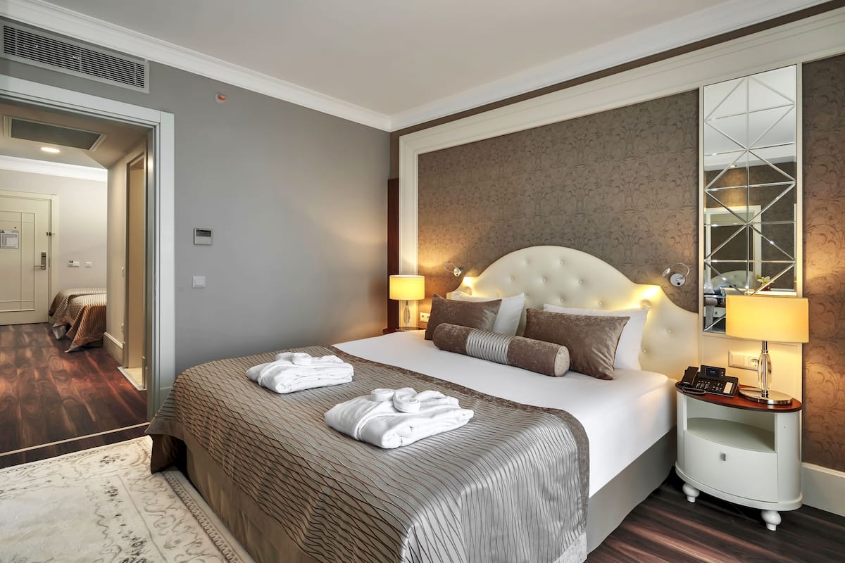 Une chambre d’hôtel avec un grand lit, deux tables de chevet avec des lampes, un miroir et une porte donnant sur une autre pièce.