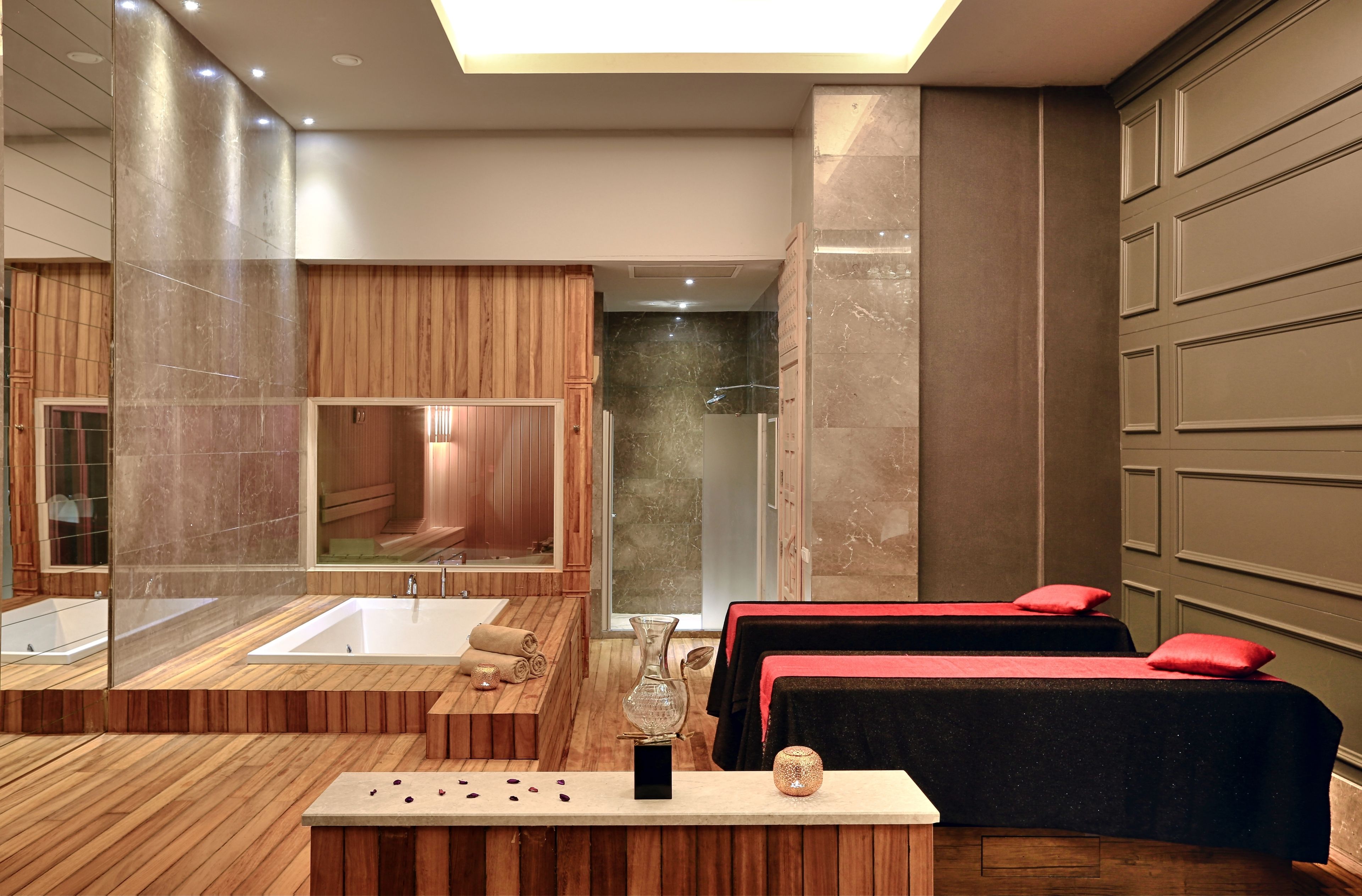 A modern spa room with a large bathtub, wooden paneling, and two massage tables with red pillows.