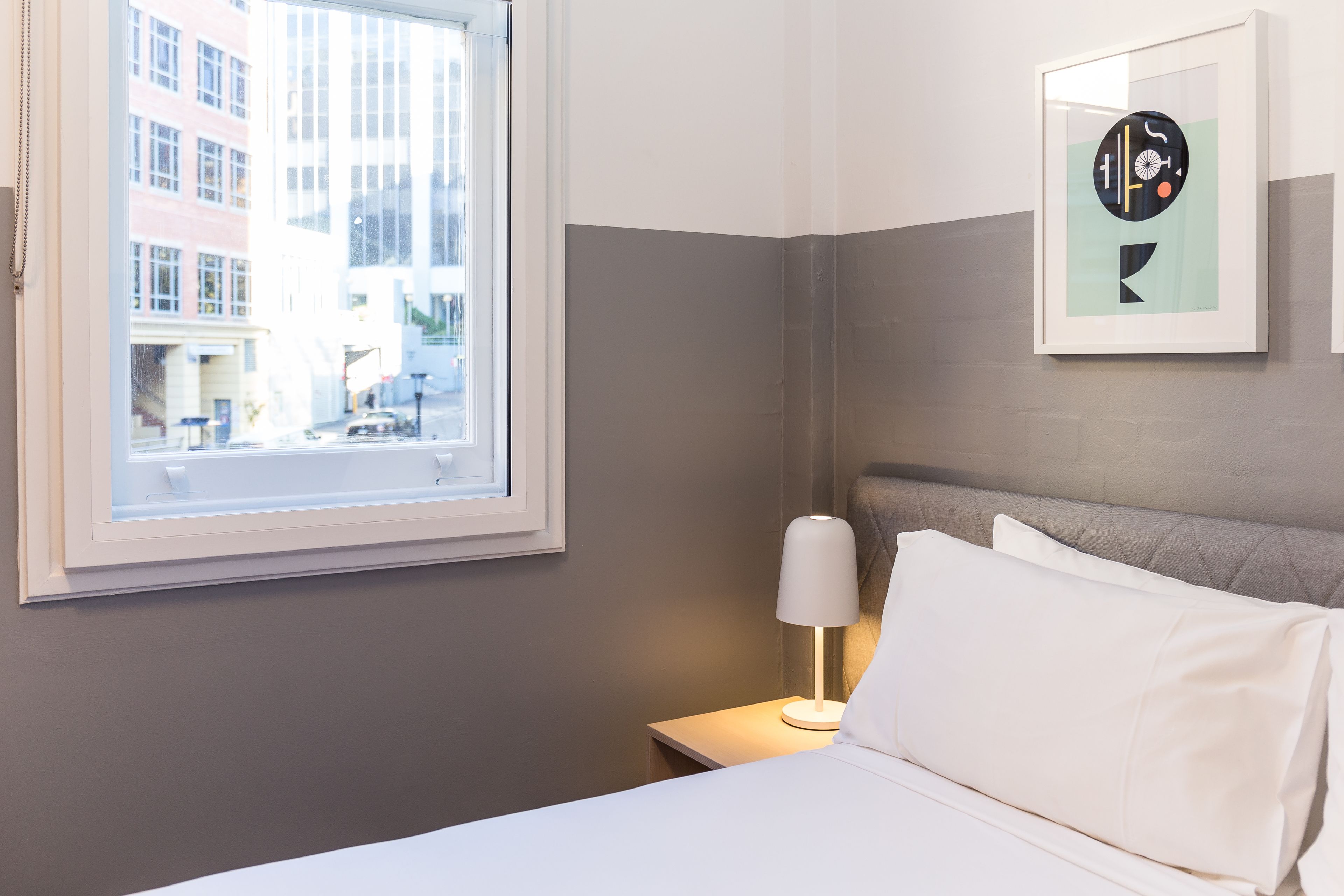 Une chambre d’hôtel avec un lit, une table de chevet avec une lampe, une œuvre d’art encadrée au mur et une fenêtre donnant sur la ville.