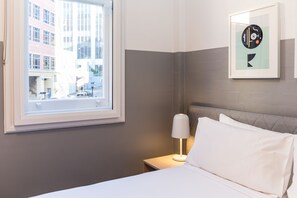 A hotel room with a bed, a nightstand with a lamp, a framed artwork on the wall, and a window with a city view.