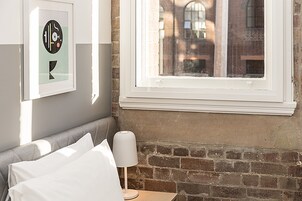 Hotellihuone, jossa on sänky, yöpöytä lampun kanssa, seinällä oleva kehystetty kuva ja ikkuna, josta on näkymä rakennuksiin.