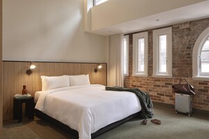 A bedroom with a bed, a nightstand, a lamp, a suitcase, and a view of a brick wall and windows.