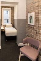 A hotel room with a bed, a chair, a nightstand, a lamp, and a window with a view of a building.