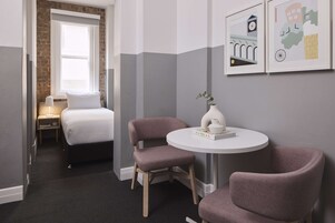 Una camera d'albergo con un letto, due sedie, un tavolino con un vaso e un libro, e una finestra con persiane.