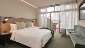 Habitación de hotel con cama, sofá, mesita de noche, lámpara y ventana con cortinas.