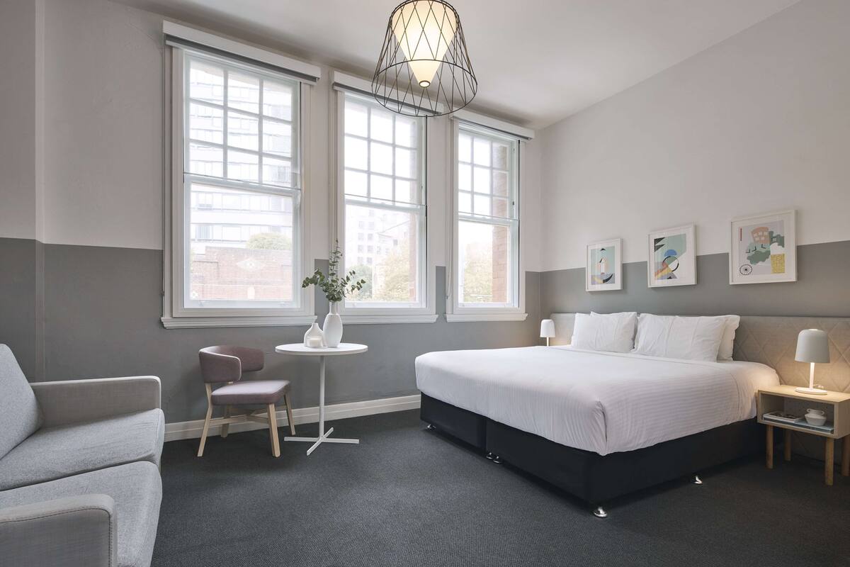 A modern hotel room with a large bed, a desk, and a chair. There are two windows providing natural light and a view of the outside.