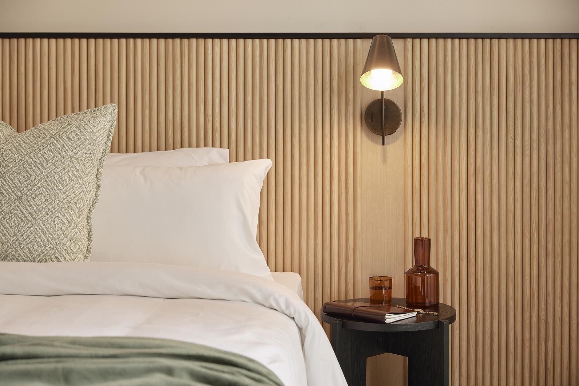 Una cama bien hecha con almohadas, una mesita de noche con lámpara y una pared con listones de madera verticales.