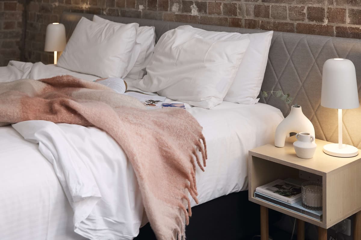 A neatly made bed with a pink blanket, white pillows, and a bedside table with a lamp and a vase.