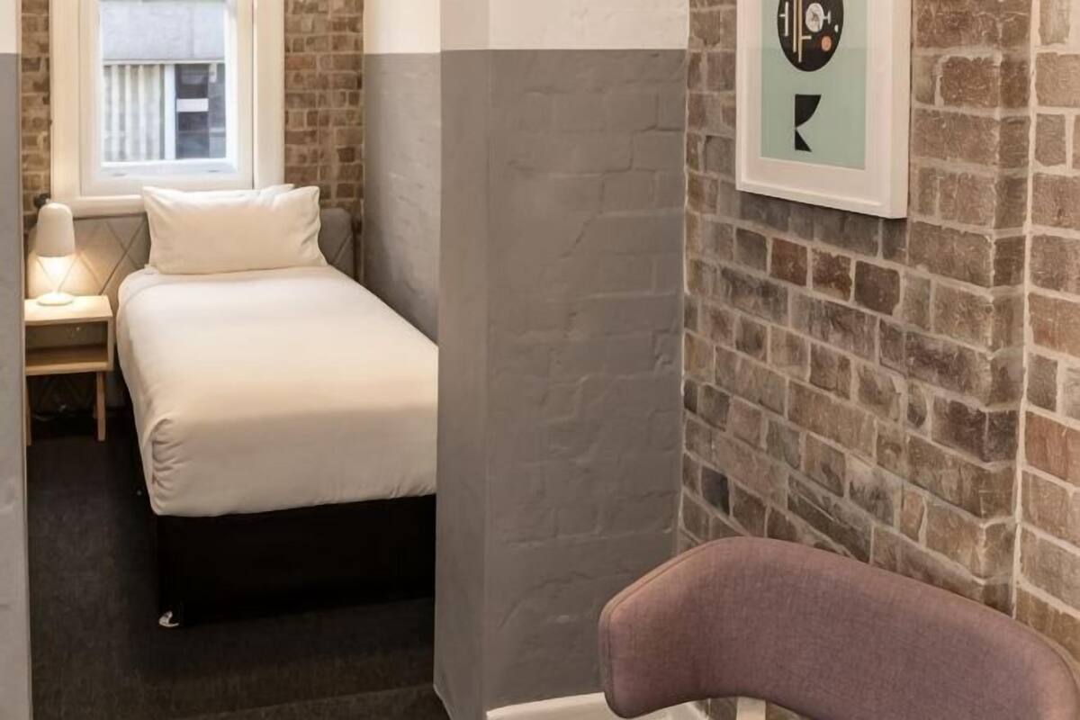 A bedroom with a bed, a chair, a window, and a brick wall.