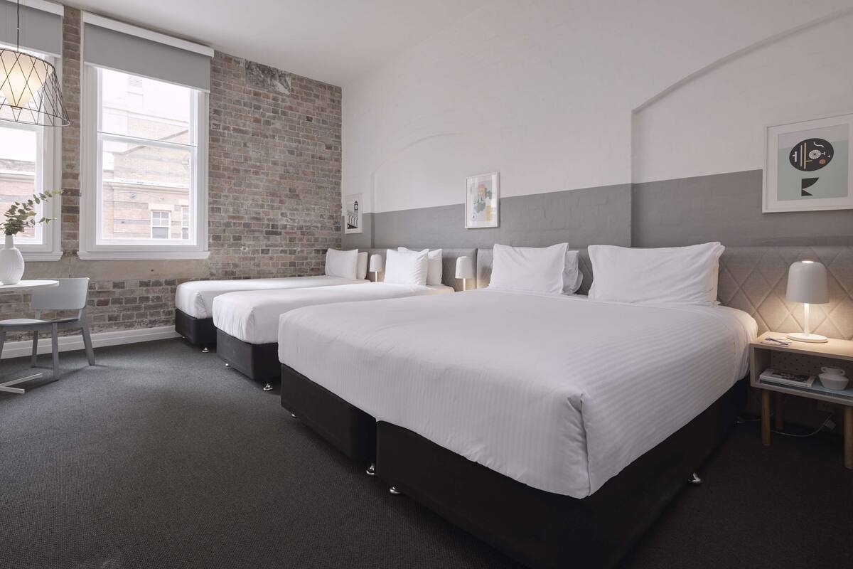 A hotel room with two beds, a desk, and a window with a view of buildings.