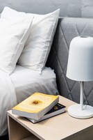 A neatly made bed with white bedding, a grey upholstered headboard, and a bedside table with a white lamp and two books.