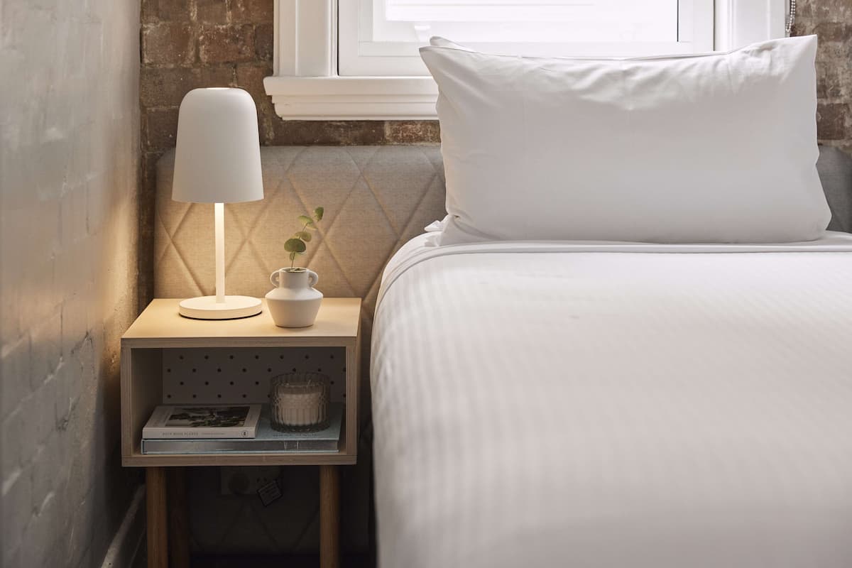 A neatly made bed with white bedding, a bedside lamp, a small potted plant, and a book on a wooden nightstand.