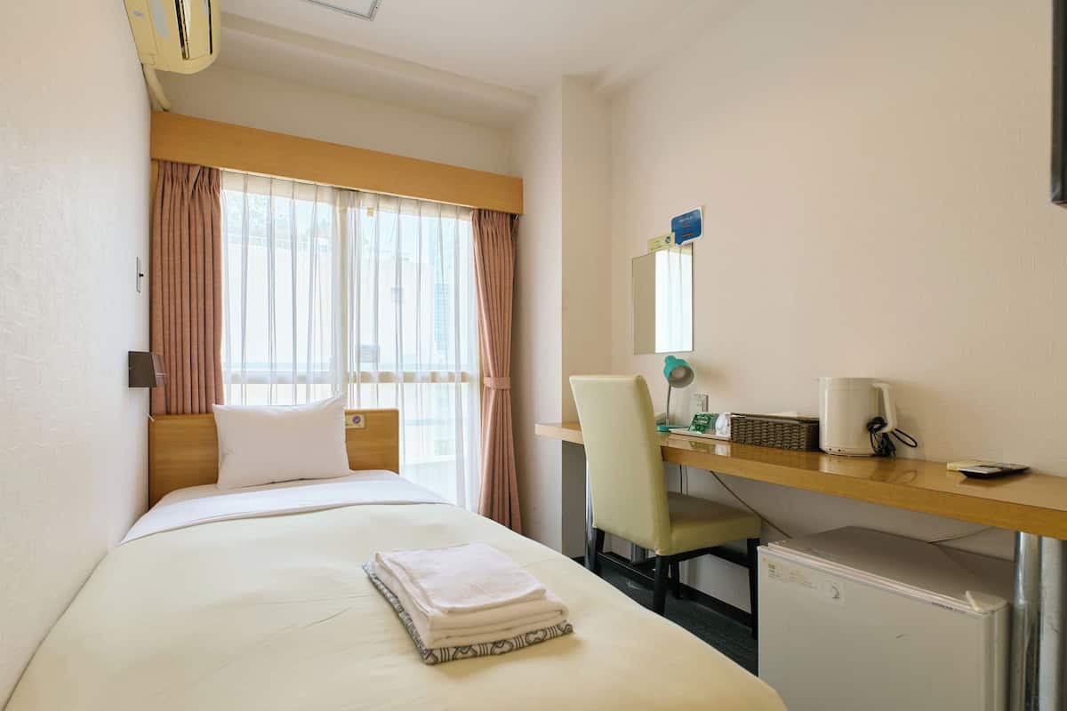 A hotel room with a bed, a desk, a chair, a small refrigerator, and a window with curtains.
